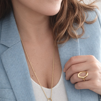 Conjunto de joias douradas com brincos ovalados, anel e colar com pendente redondo em mulher com blazer azul