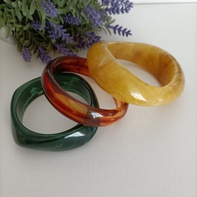 Três pulseiras de resina coloridas ao lado de flores lilás artificiais.