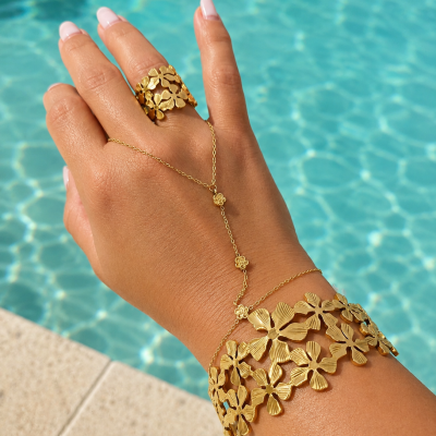 Mão com anel, pulseira larga e fina dourados com padrão de flores perto de piscina