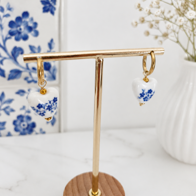 Brincos dourados com pendentes em coração branco com padrão floral azul num suporte dourado com base de madeira