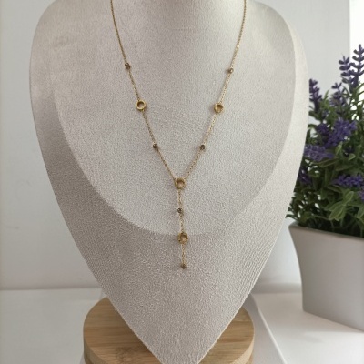 Colar dourado com círculos em busto bege e planta roxa ao fundo