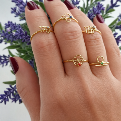 Mão com cinco anéis dourados de símbolos zodiacais sobre fundo de lavanda roxa