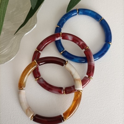 Três pulseiras de resina colorida em azul, vermelho, branco e âmbar com anéis dourados, sobre tecido branco e folhas verdes.