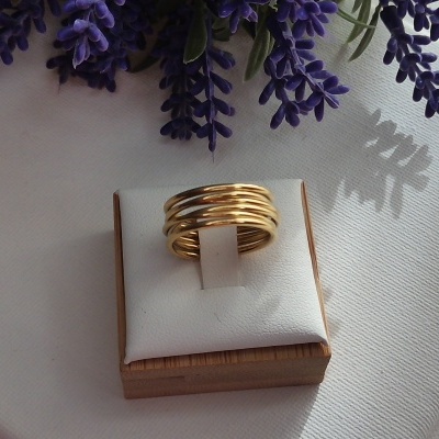 Anel dourado com fios entrelaçados sobre almofada branca e base de madeira, flores roxas ao fundo