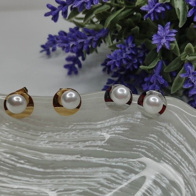 Brincos de botão com pérolas, bases dourada e prateada, sobre superfície branca texturada e flores roxas