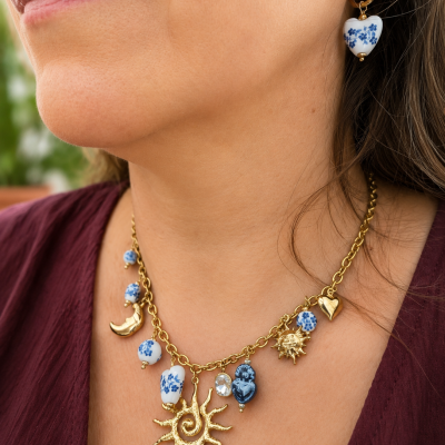 Colar dourado com pendentes em porcelana branca e azul floral e elementos em forma de sol, coração e lua junto com brinco de argola dourada com pendente de coração.