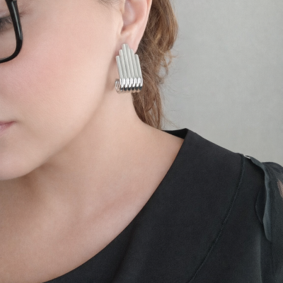 Brincos de metal prateado com design em camadas usados por mulher com blusa preta e óculos pretos.