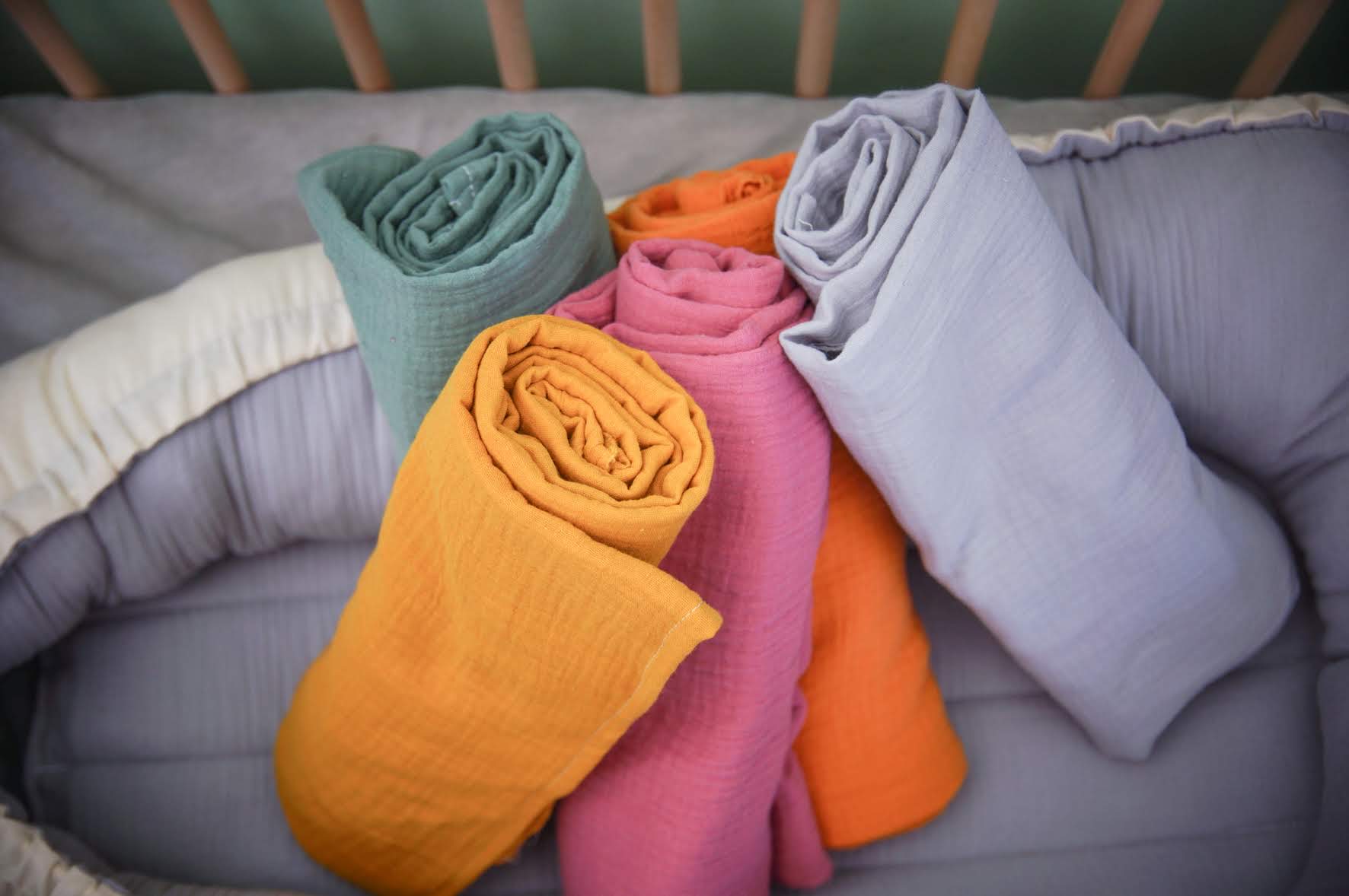 Ninho Cru Lençóis de algodão enrolados em cores verde, amarelo, rosa e cinza numa cama cinzenta