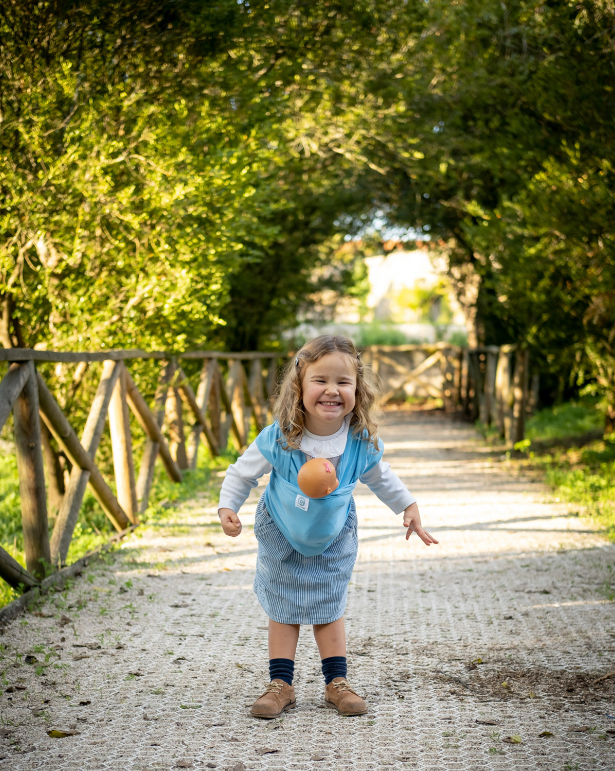 ISling4kid - Porta-bonecas Criança com sling azul claro ao ar livre num caminho com cerca de madeira e árvores verdes