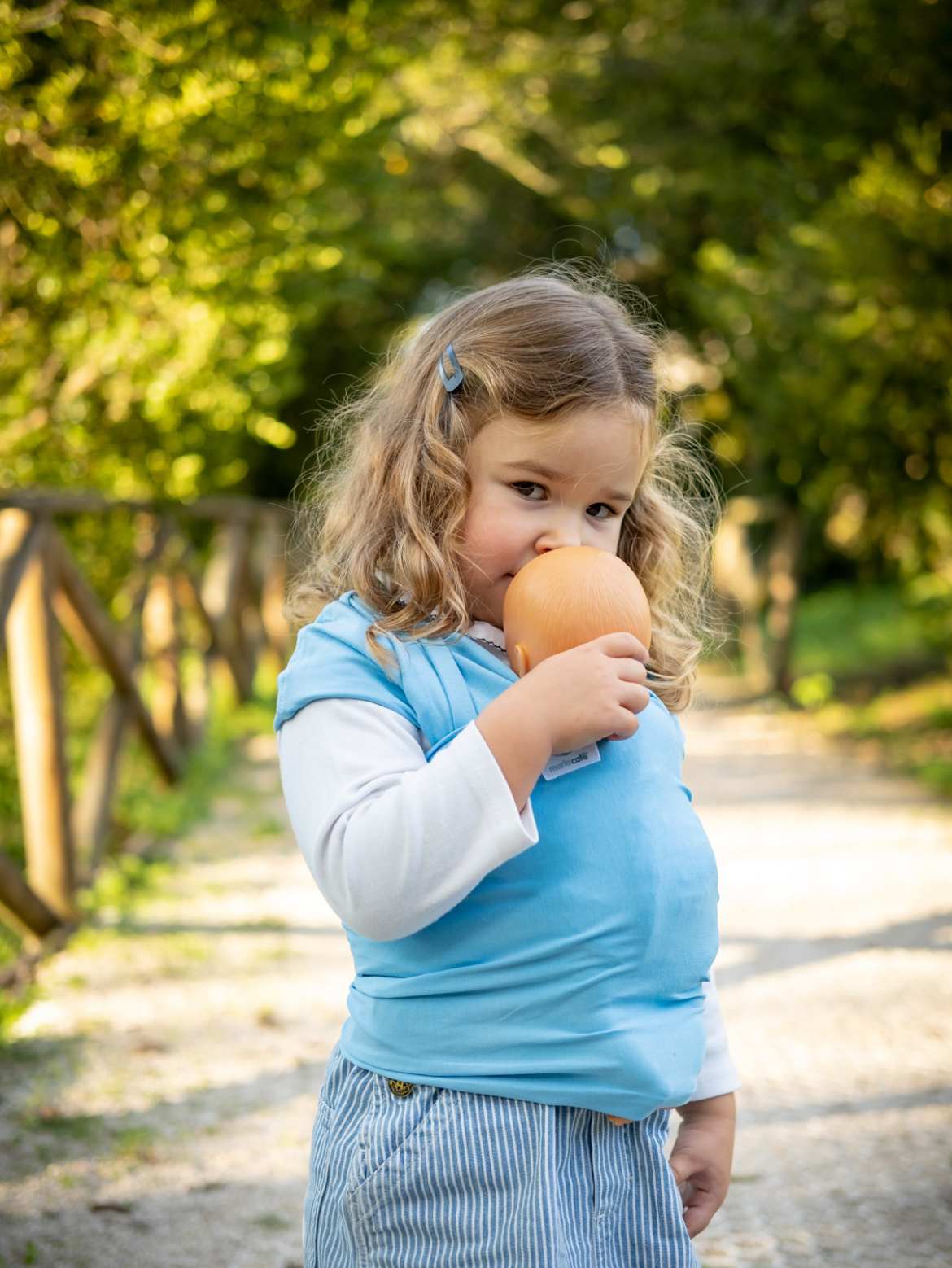 ISling4kid - Porta-bonecas Criança loira a cheirar laranja ao ar livre, vestindo colete azul e calças riscas.