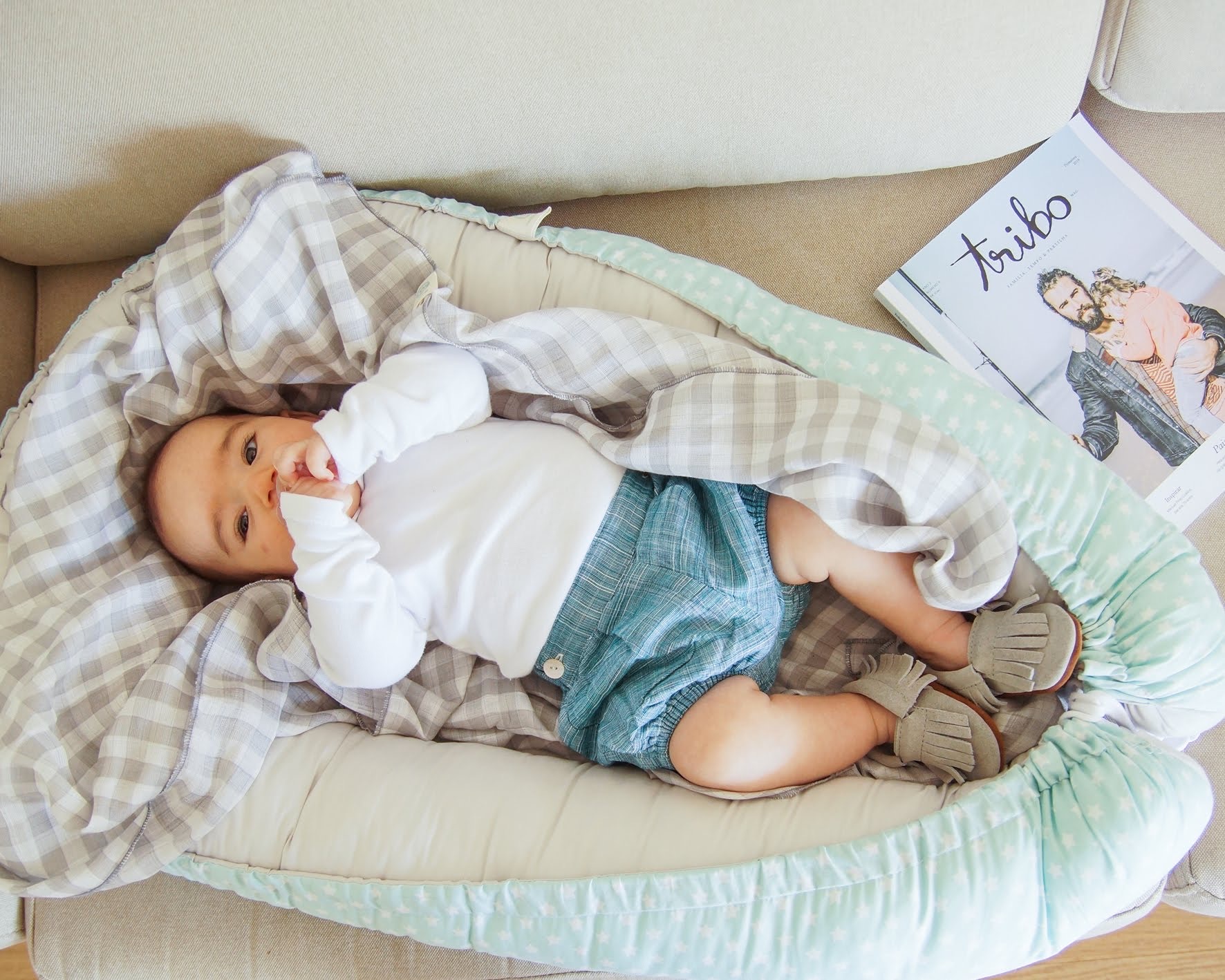 Ninho liso com padrão Bebé deitado em cama portátil com coberta xadrez ao lado de revista Tribo num sofá bege.
