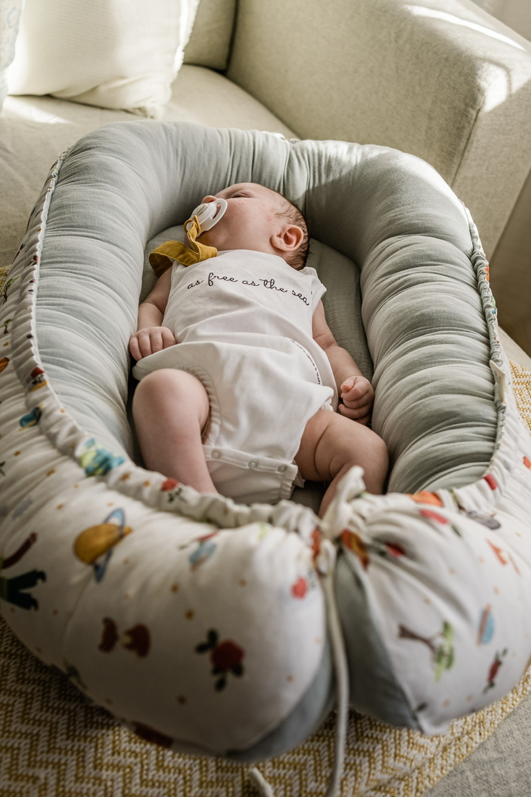 Ninho Principezinho Bebé a dormir em almofada ninho com padrão colorido e interior cinzento sobre poltrona beige