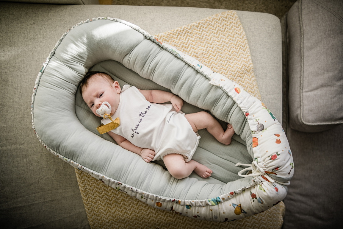 Ninho Principezinho Berço portátil acolchoado com bebé dentro, colocado sobre manta bege e sofá cinza