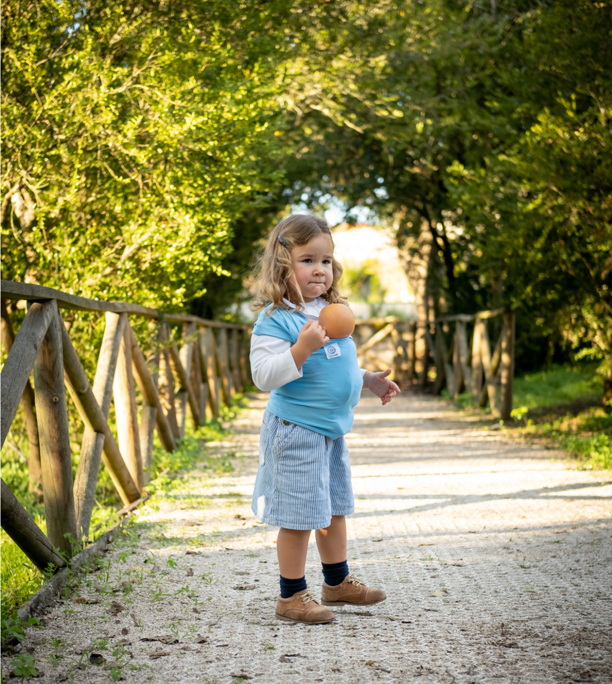 ISling4kid - Porta-bonecas Criança com suporte para bebé azul e calções cinzentos num caminho exterior
