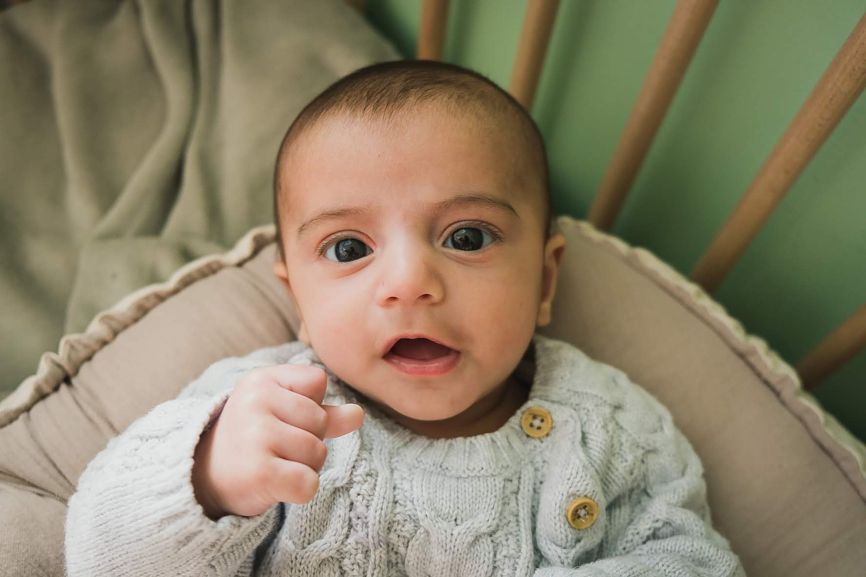 Ninho Cru Bebé com camisola cinzenta de malha sentado numa almofada creme