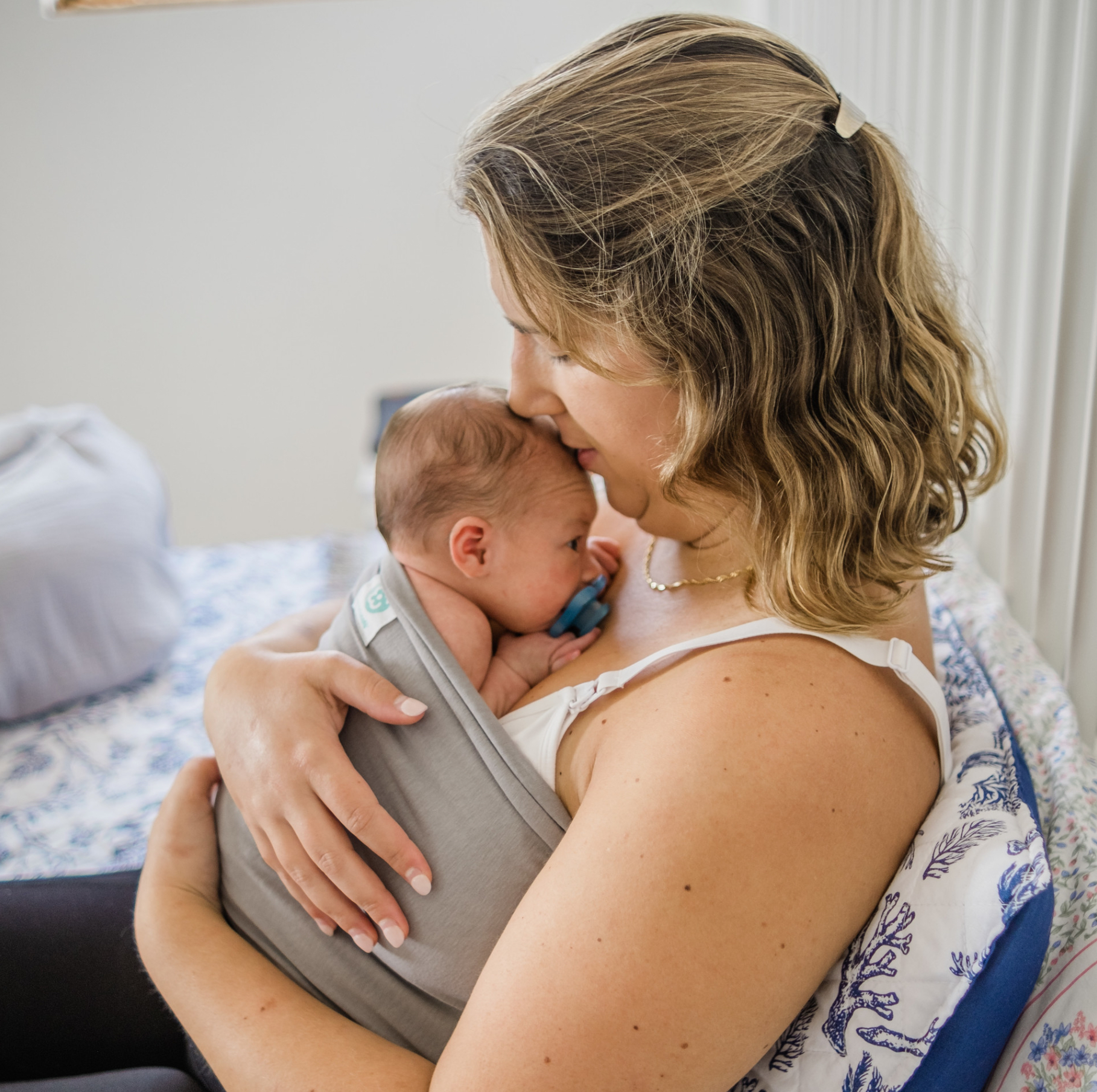 IGaroo Mulher a segurar bebé recém-nascido num sling cinzento com chupeta azul, sentada numa cadeira com almofadas brancas e azuis.