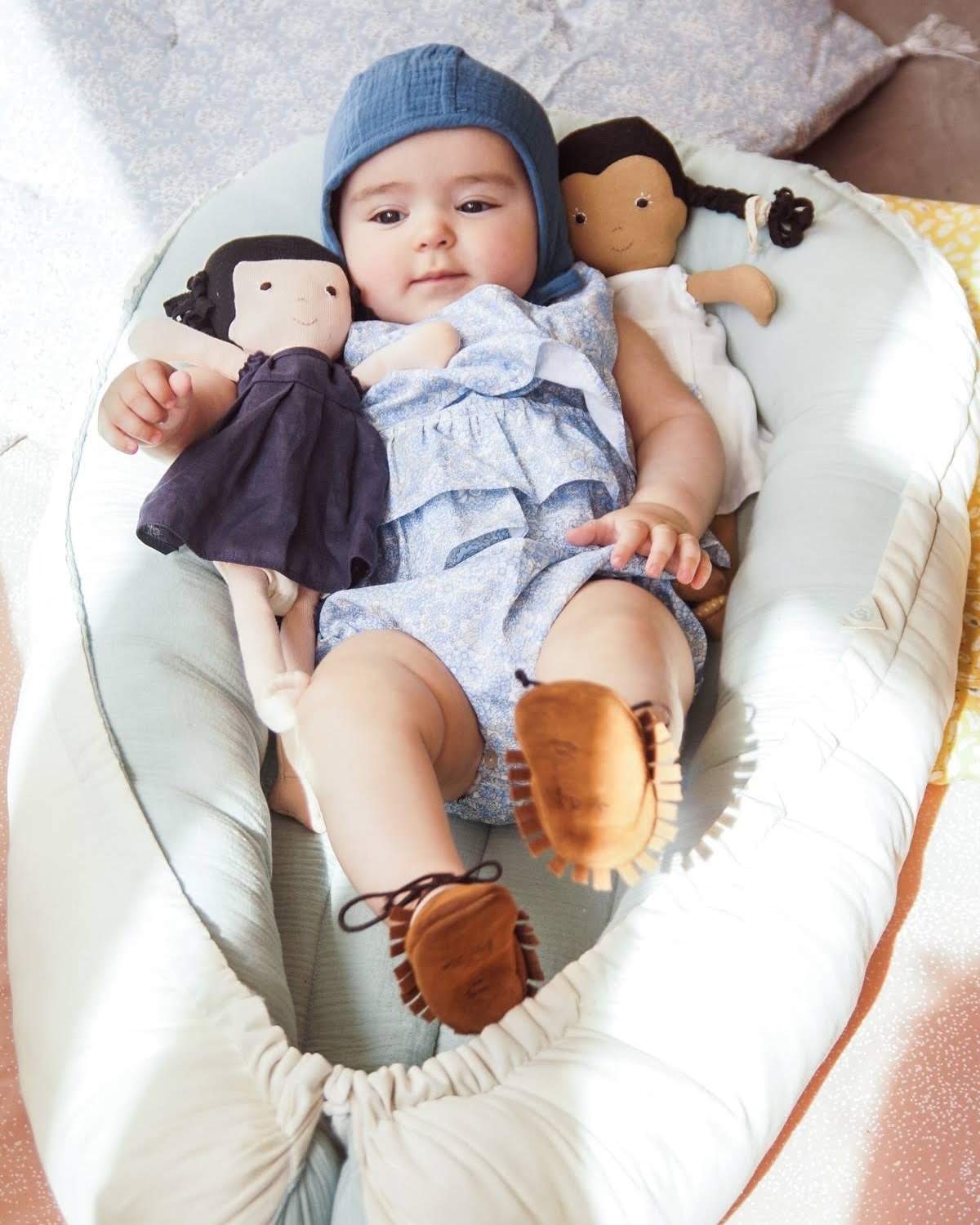 Ninho Cru Bebé deitada em almofada segurando duas bonecas de tecido