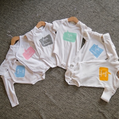 Cinco bodies brancos para bebé com quadrados coloridos e texto em inglês no peito, pendurados em cabides de madeira