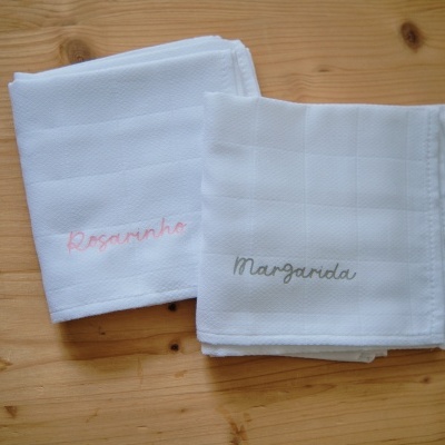 Dois panos de cozinha brancos sobre mesa de madeira, com nomes bordados