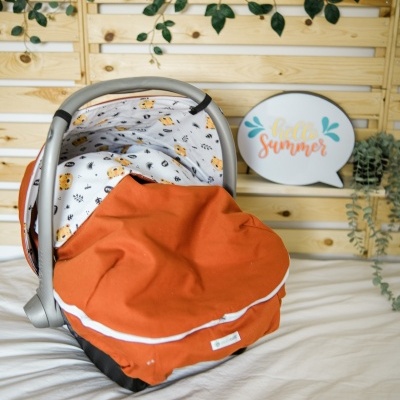 Cadeira de bebé para carro com tecido interior branco com desenhos de animais e exterior laranja, sobre cama branca e painel de madeira ao fundo.