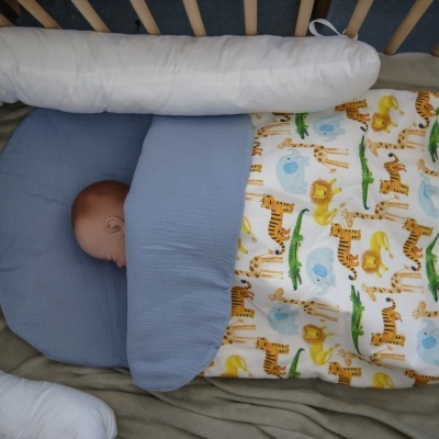 Bebé num saco de dormir infantil com padrão de animais, numa cama com cabeceira protetora branca.