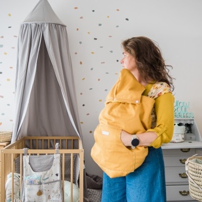 Mulher com bebé em saco de inverno amarelo num quarto de bebé com berço de madeira e dossel cinzento