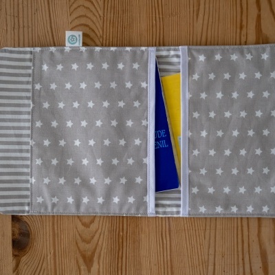Estojo de tecido cinza com estrelas brancas e interior riscado, com livro azul e amarelo dentro, sobre chão de madeira.
