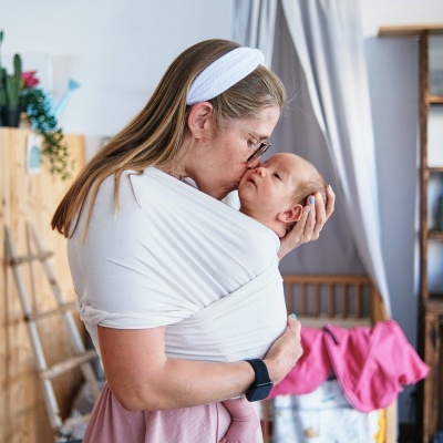 Mulher com bebé numa faixa de tecido branco num quarto com cortinas cinza e mobília de madeira