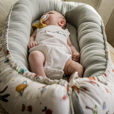 Bebé a dormir em almofada ninho com padrão colorido e interior cinzento sobre poltrona beige
