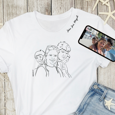 T-shirt branca com desenho linear de três rostos e texto junto à gola, smartphone com foto e roupa azul clara sobre fundo de madeira clara