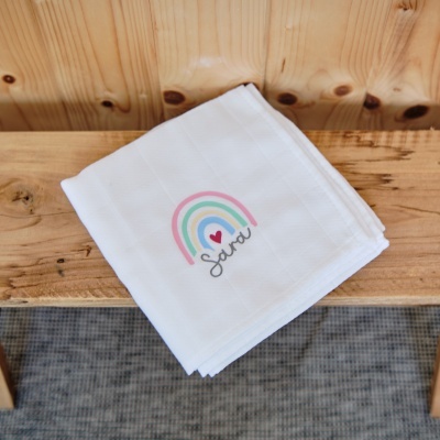 Fralda branca com arco-íris colorido e nome Sara sobre banco de madeira