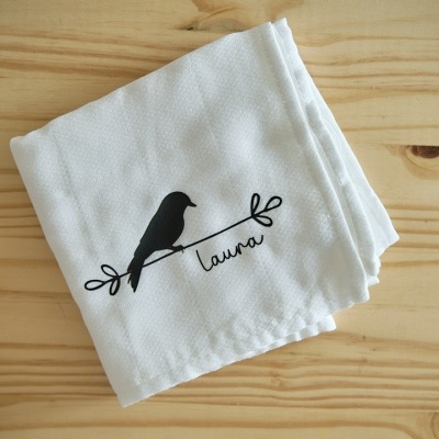 Pano branco com estampa de pássaro e nome Laura sobre mesa de madeira