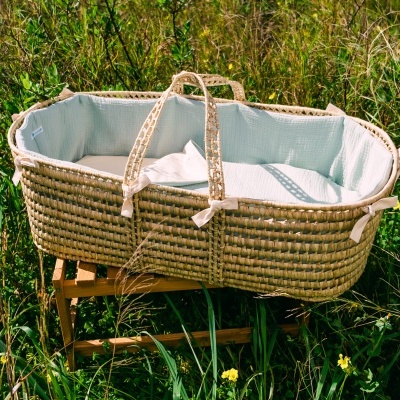 Berço de bebé em vime natural com forro branco sobre estrutura de madeira em campo.