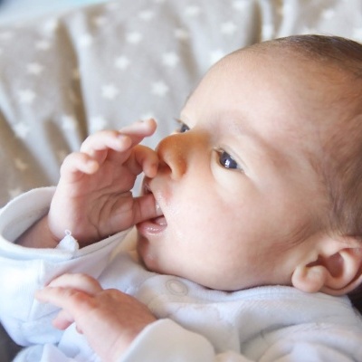 Bebé vestido de branco a morder o dedo, deitado numa almofada cinza com estrelas brancas