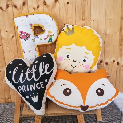 Quatro almofadas decorativas coloridas apoiadas num banco de madeira, incluindo uma almofada 'P', uma almofada coração com texto 'little PRINCE', uma almofada raposa e uma almofada de rosto de príncipe.