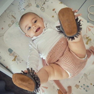 Bebé deitado com roupa clara e sapatinhos castanhos com franjas num colchão estampado