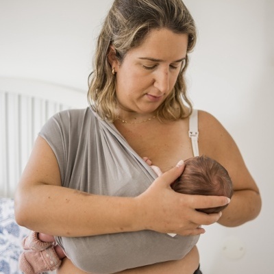 Mulher a usar sling cinzento a segurar bebé num quarto com cama branca.