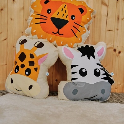 Três almofadas de pelúcia com desenhos de leão, girafa e zebra sobre superfície clara e fundo de madeira