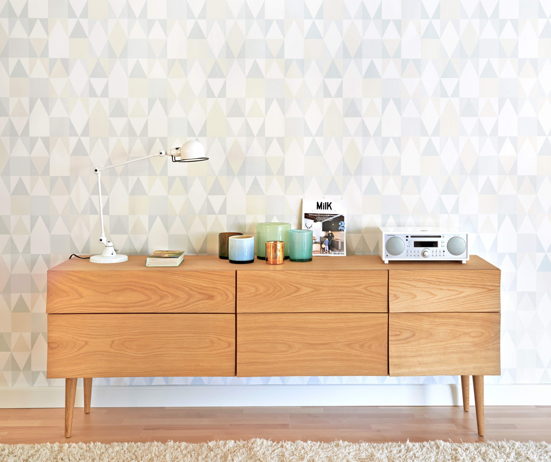 Aparador de madeira clara com lâmpada, velas, livro, revista MIK, aparelho de som branco e papel de parede geométrico