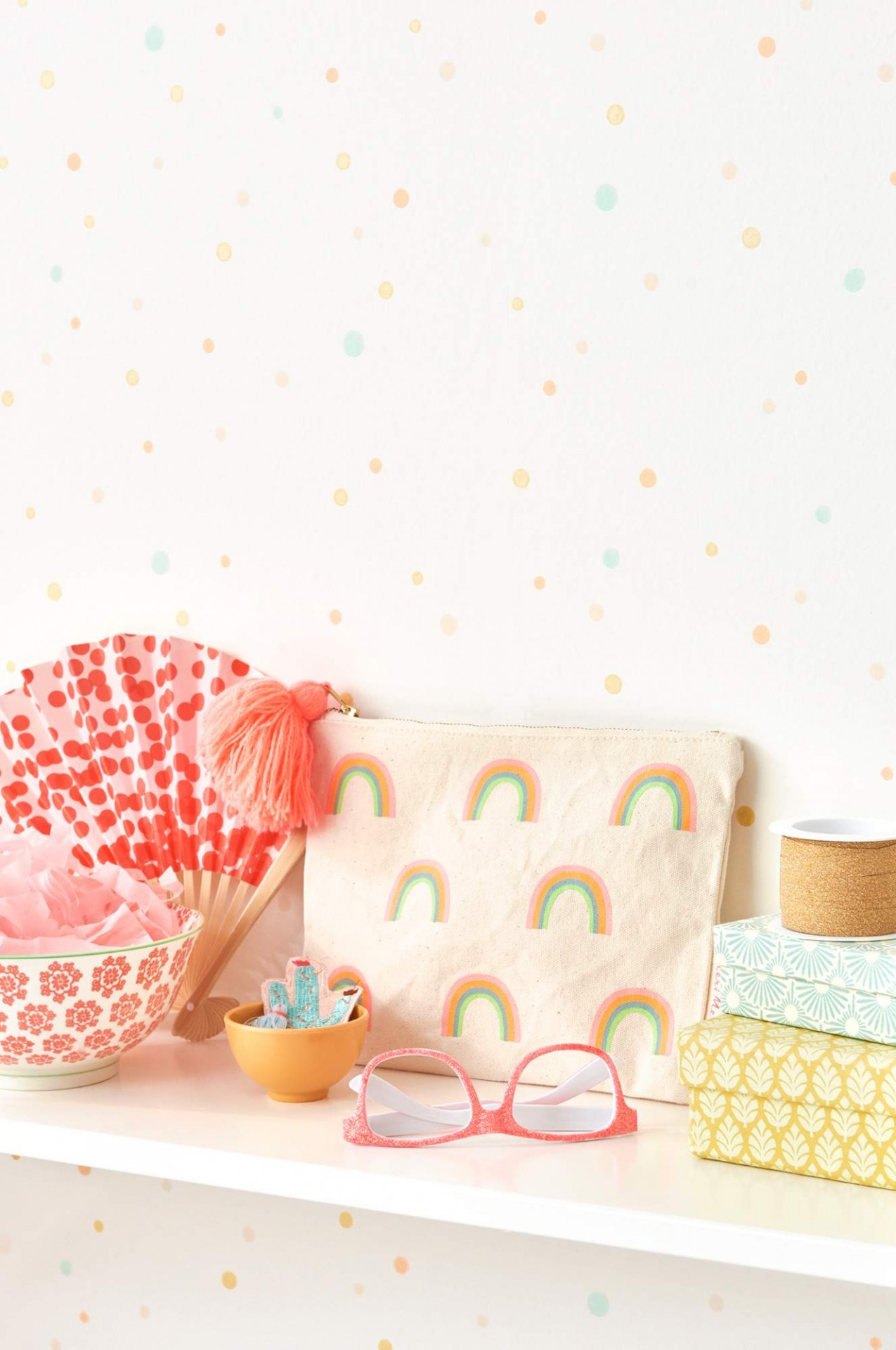 bolsa de tecido com padrão de arco-íris, leque rosa com bolinhas vermelhas, óculos cor-de-rosa, tigela branca com padrão floral vermelho, taça amarela com objetos, três caixinhas decorativas empilhadas