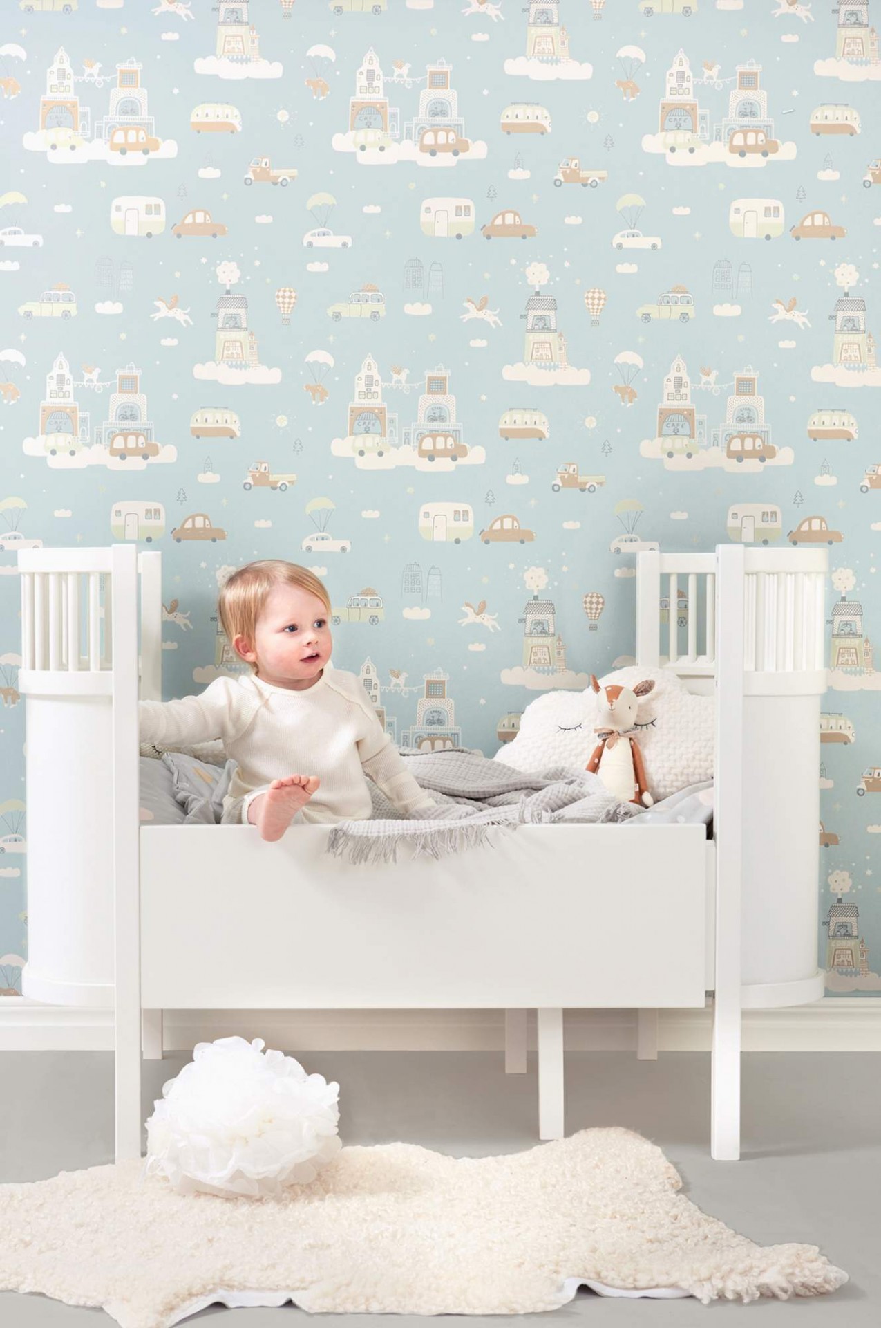Berço branco com bebé e papel de parede azul claro com desenhos de carros e faróis