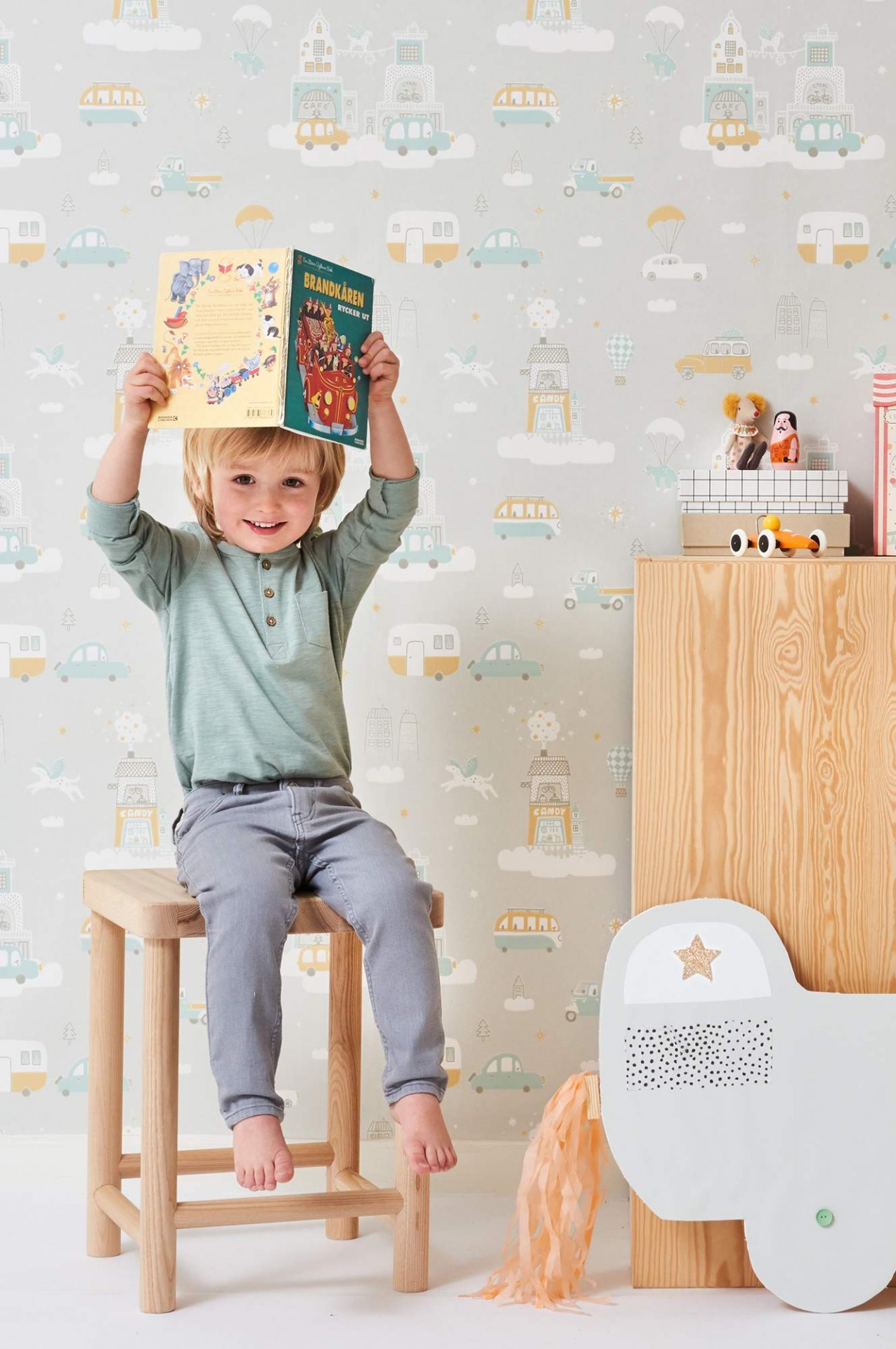 Criança sentada num banco de madeira segurando um livro colorido em quarto infantil com papel de parede pastel e móveis de madeira clara