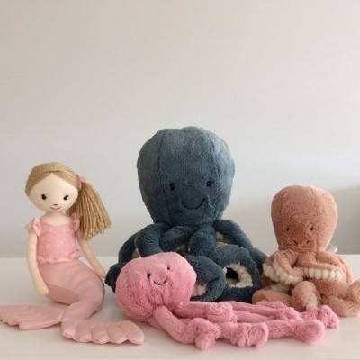 Quatro peluches: sereia rosa, polvo cinza escuro, polvo cor-de-rosa e polvo salmão numa superfície branca