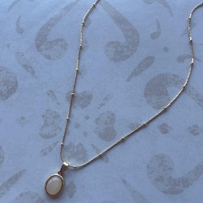 Colar dourado com pendente oval branco em fundo claro com padrão cinzento