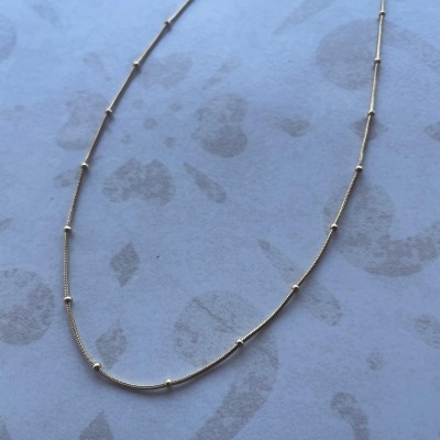 Colar fino dourado com pequenos espaçadores redondos sobre fundo azul claro com manchas escuras