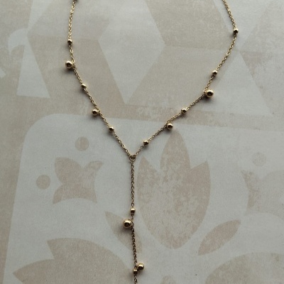 Colar delicado dourado com pequenas esferas e pendente em Y sobre fundo com padrão floral