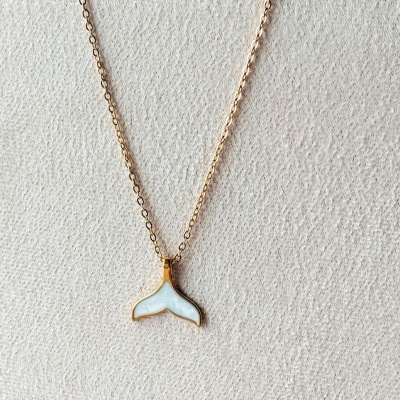 Colar dourado com pendente branco em forma de cauda de baleia sobre fundo branco texturizado