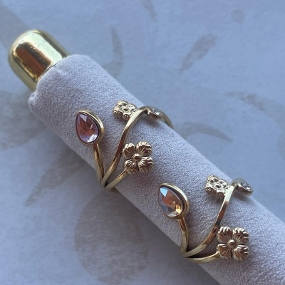 Anel dourado com flores e pedras rosa em suporte de tecido cinzento.
