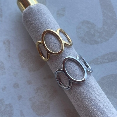 Dois anéis ovais em dourado e prata sobre suporte de tecido cinza claro.