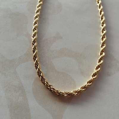 Colar dourado com corrente trançada em fundo bege com padrão abstrato.